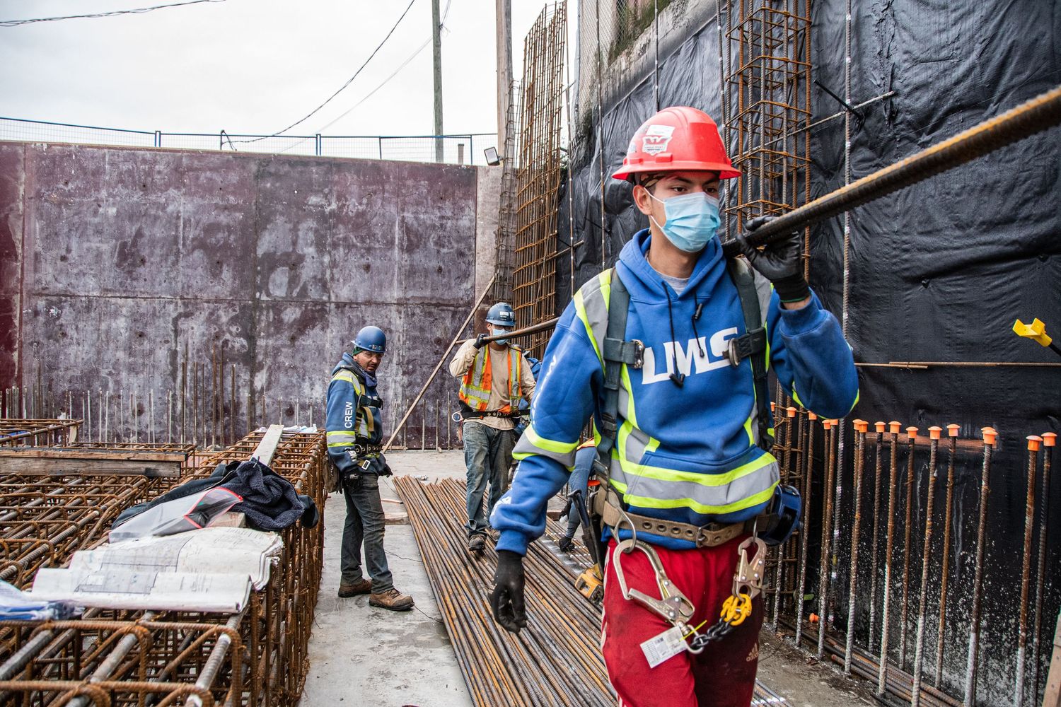 Construction workers in safety gear and masks work amid steel rebar at a building site.