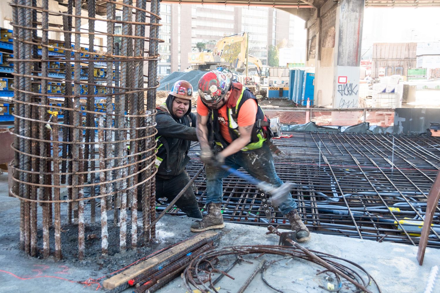 Two construction workers in hard hats tying rebar at an urban building foundation site.