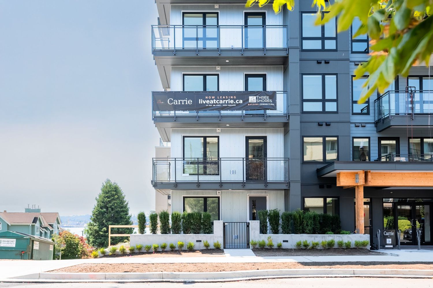 Modern multi-story residential condominium building with black frame windows, balconies, and real estate signage on a sunny day.