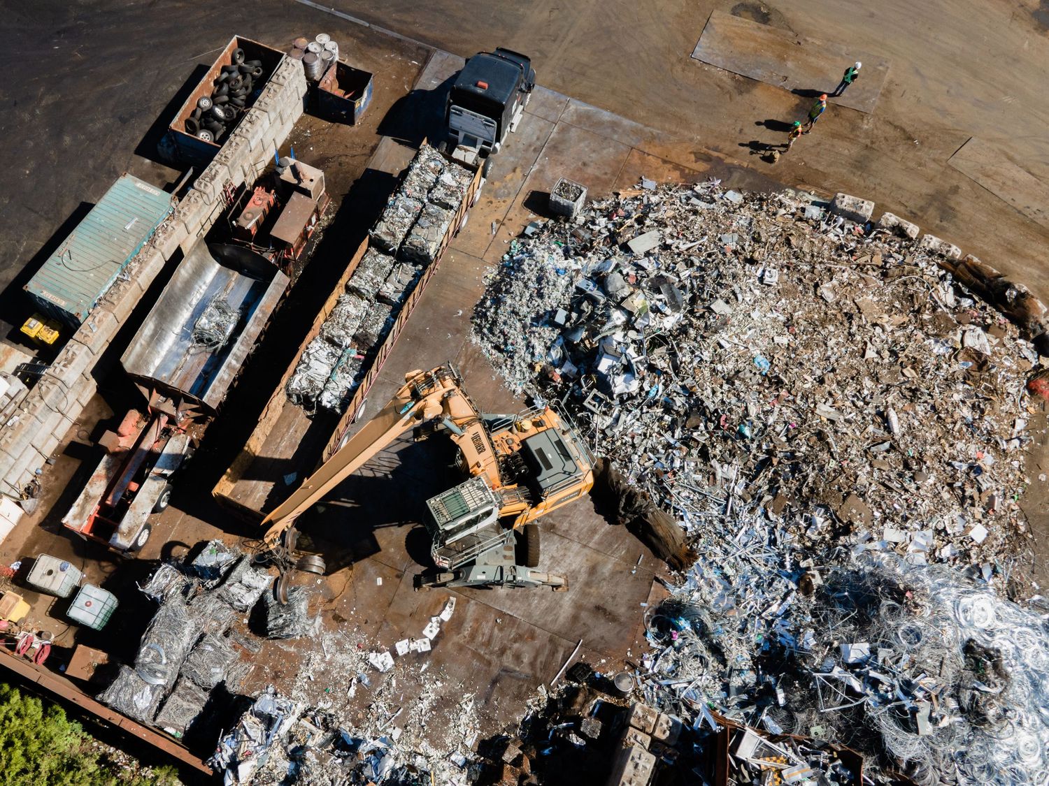 Aerial drone view of scrapyard with excavator machinery sorting through large pile of metal debris.