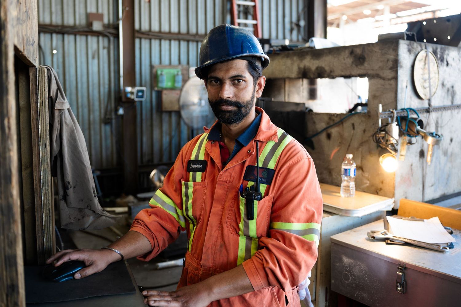 Male construction worker in orange safety coveralls and blue hard hat posing in industrial workshop.