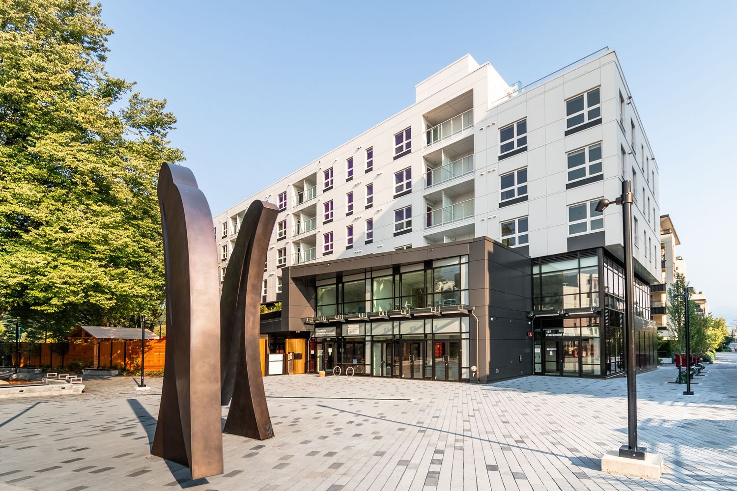 Modern white and dark mixed-use residential building with public plaza, sculpture, and surrounding mature trees.