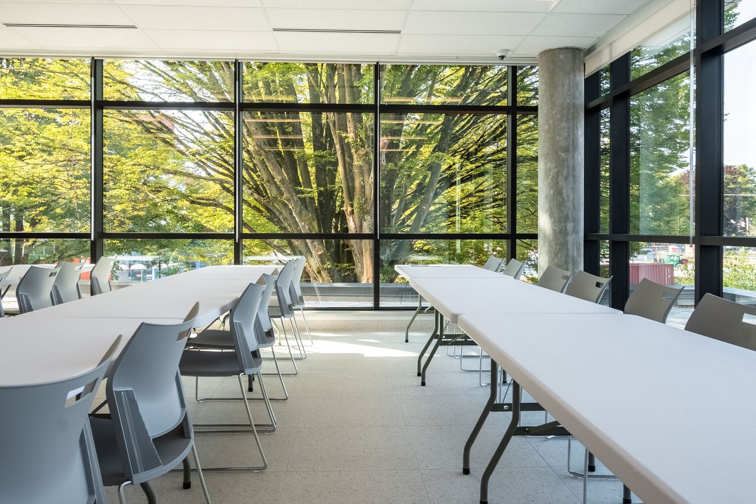 Bright modern conference room with long white tables, gray chairs, and floor-to-ceiling windows overlooking green trees.