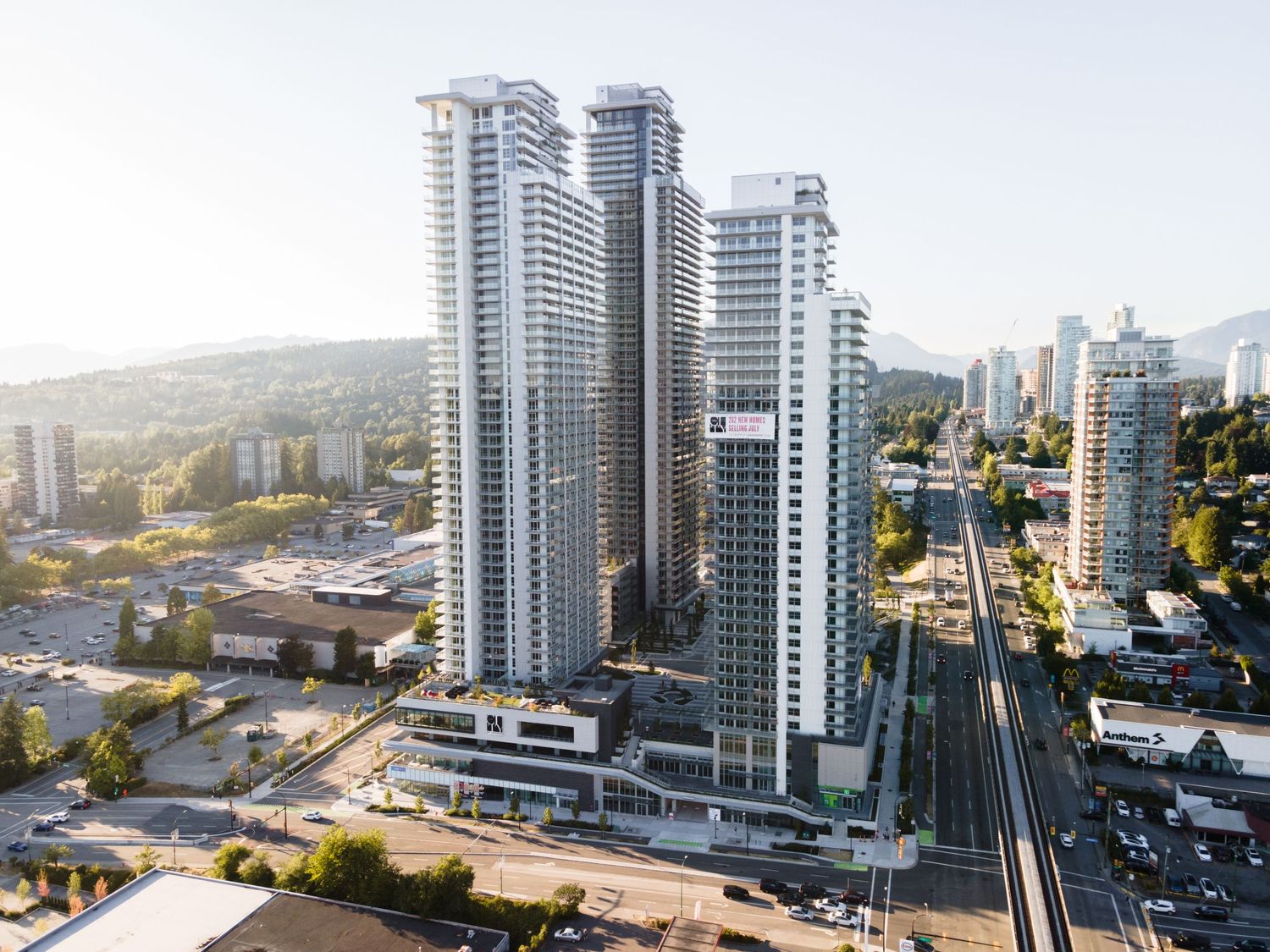 Aerial view of The City of Lougheed residential towers beside SkyTrain tracks with mountain views.