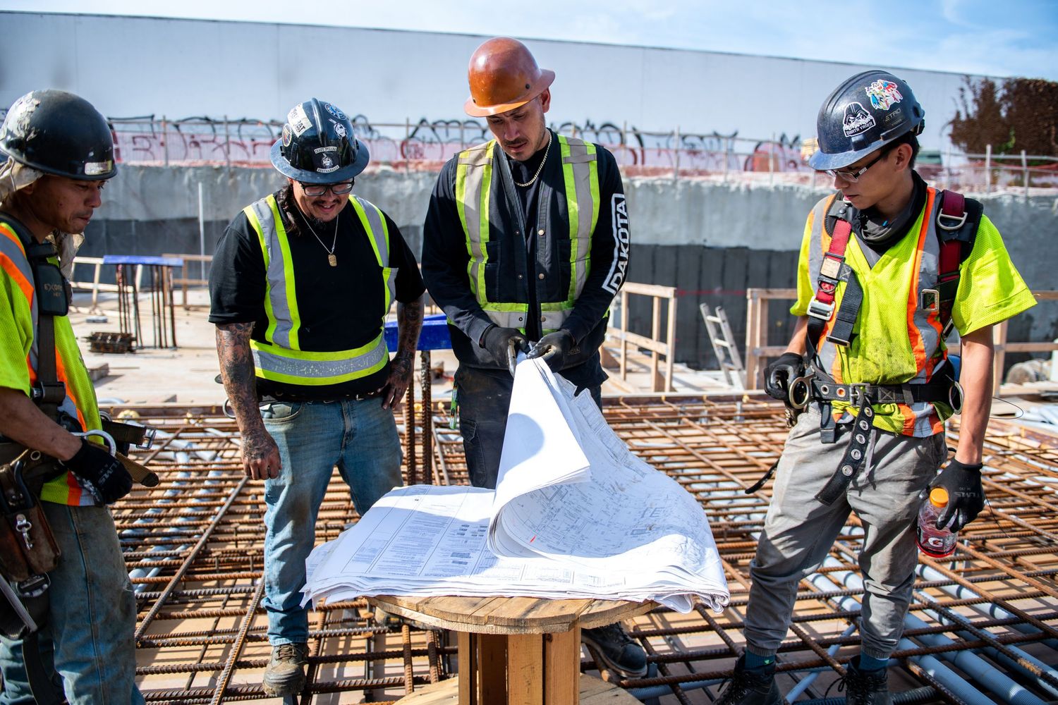 Construction workers in safety gear reviewing blueprints at an active building site with rebar.