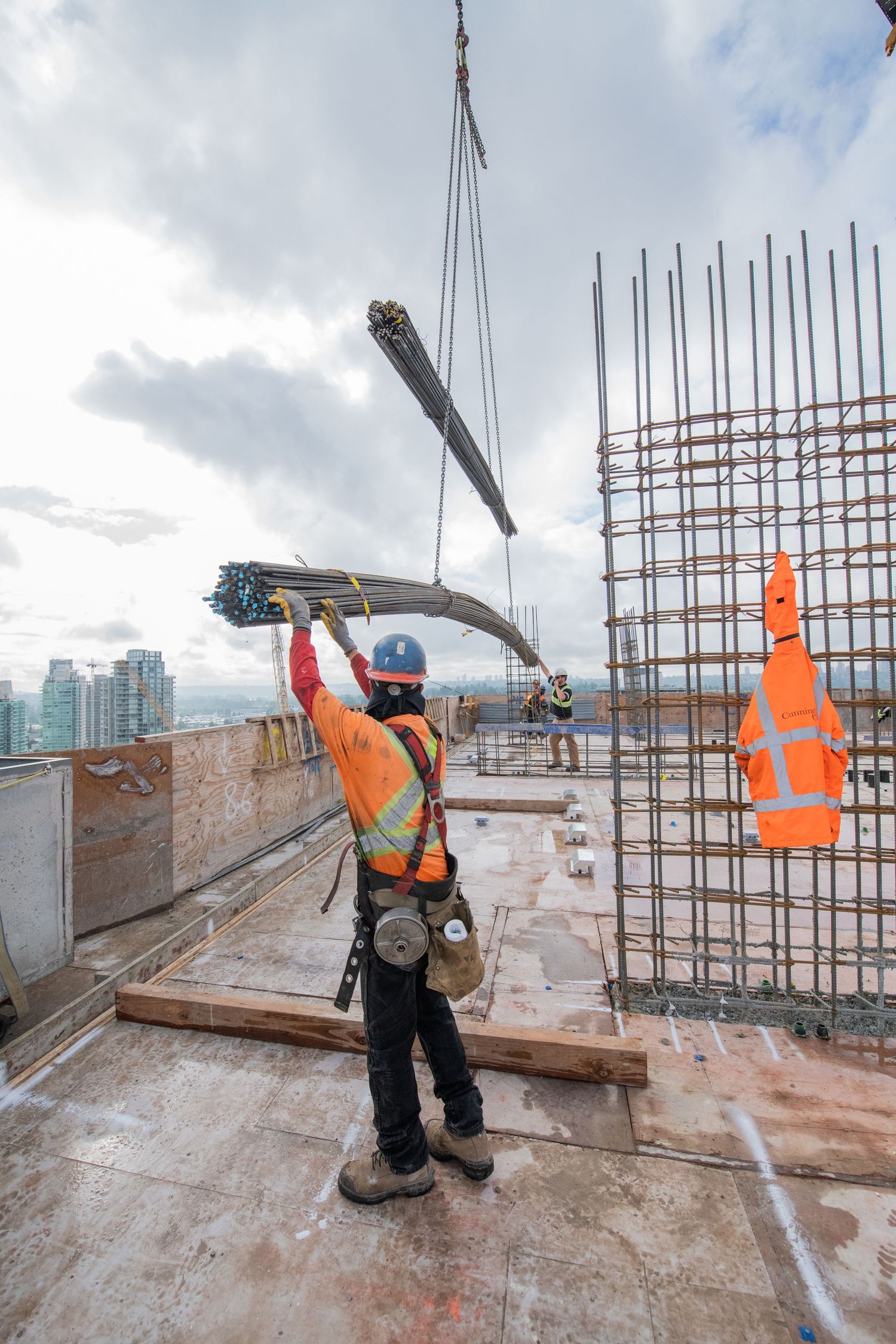 Construction worker guides a crane lifting steel rebar on a high-rise building rooftop with city views.