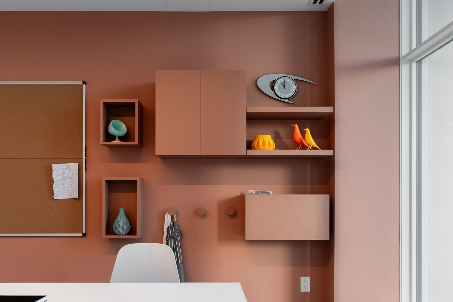 Terracotta-toned office wall with floating shelves displaying colorful decorative objects above a white desk.
