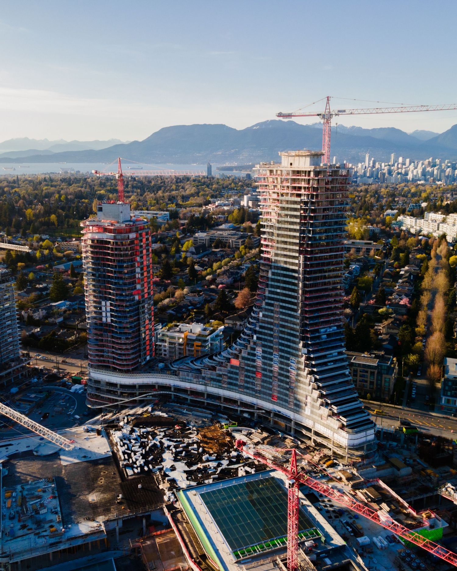 Aerial view of two high-rise towers under construction with cranes against a mountain backdrop in Vancouver.