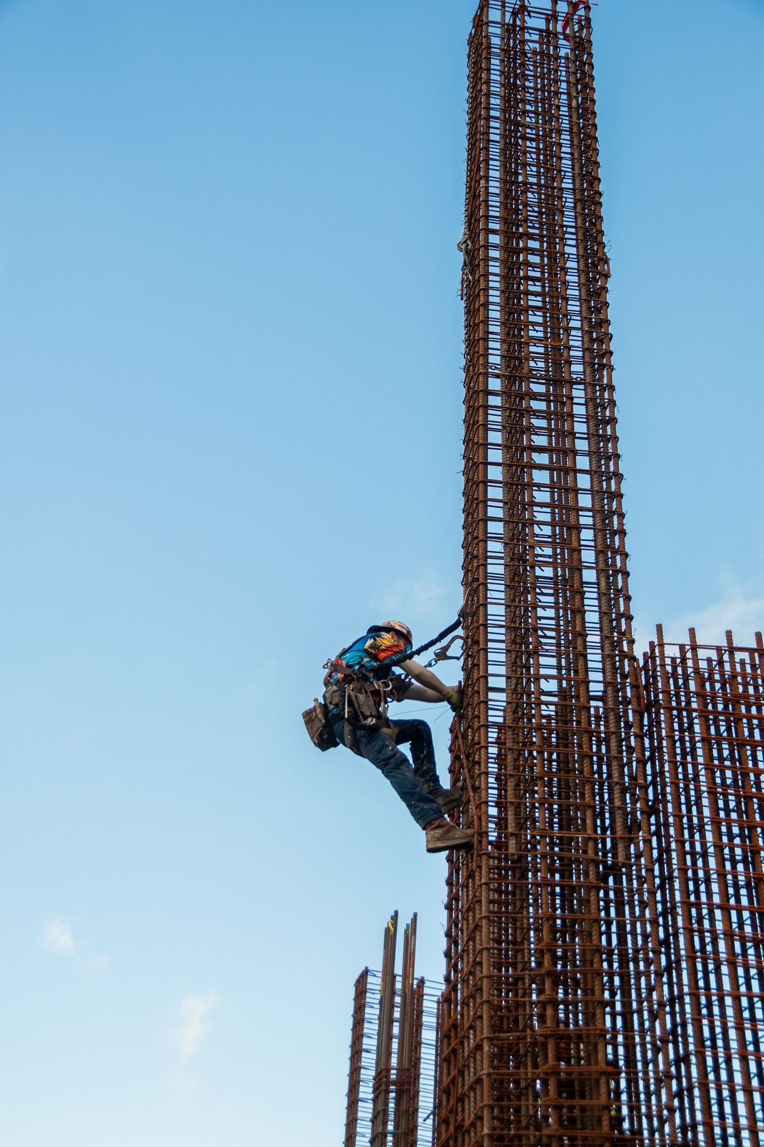 Construction worker climbing tall rebar column structure against clear blue sky on building site.