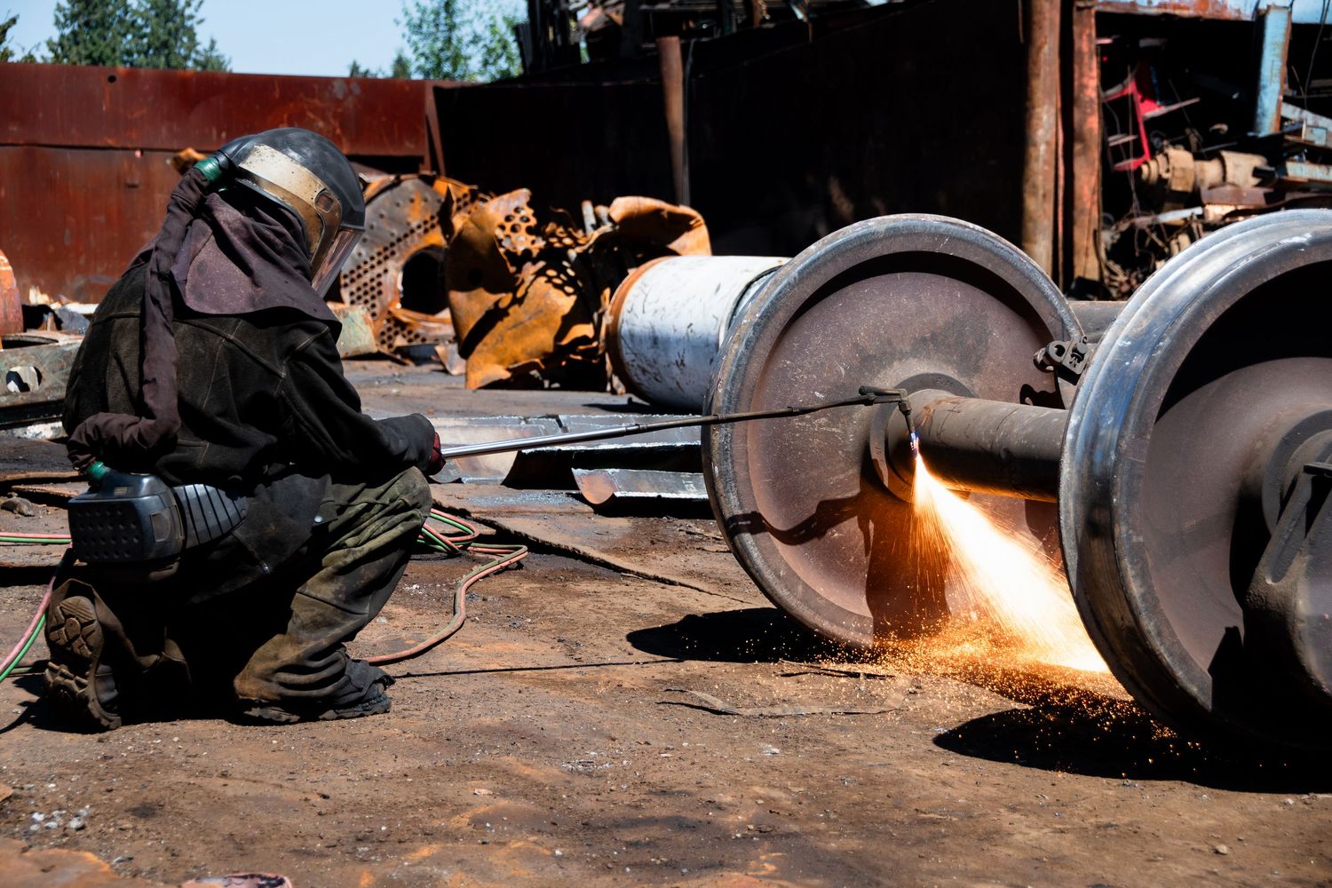 Worker in protective gear using cutting torch on large metal wheel axle outdoors at scrapyard.