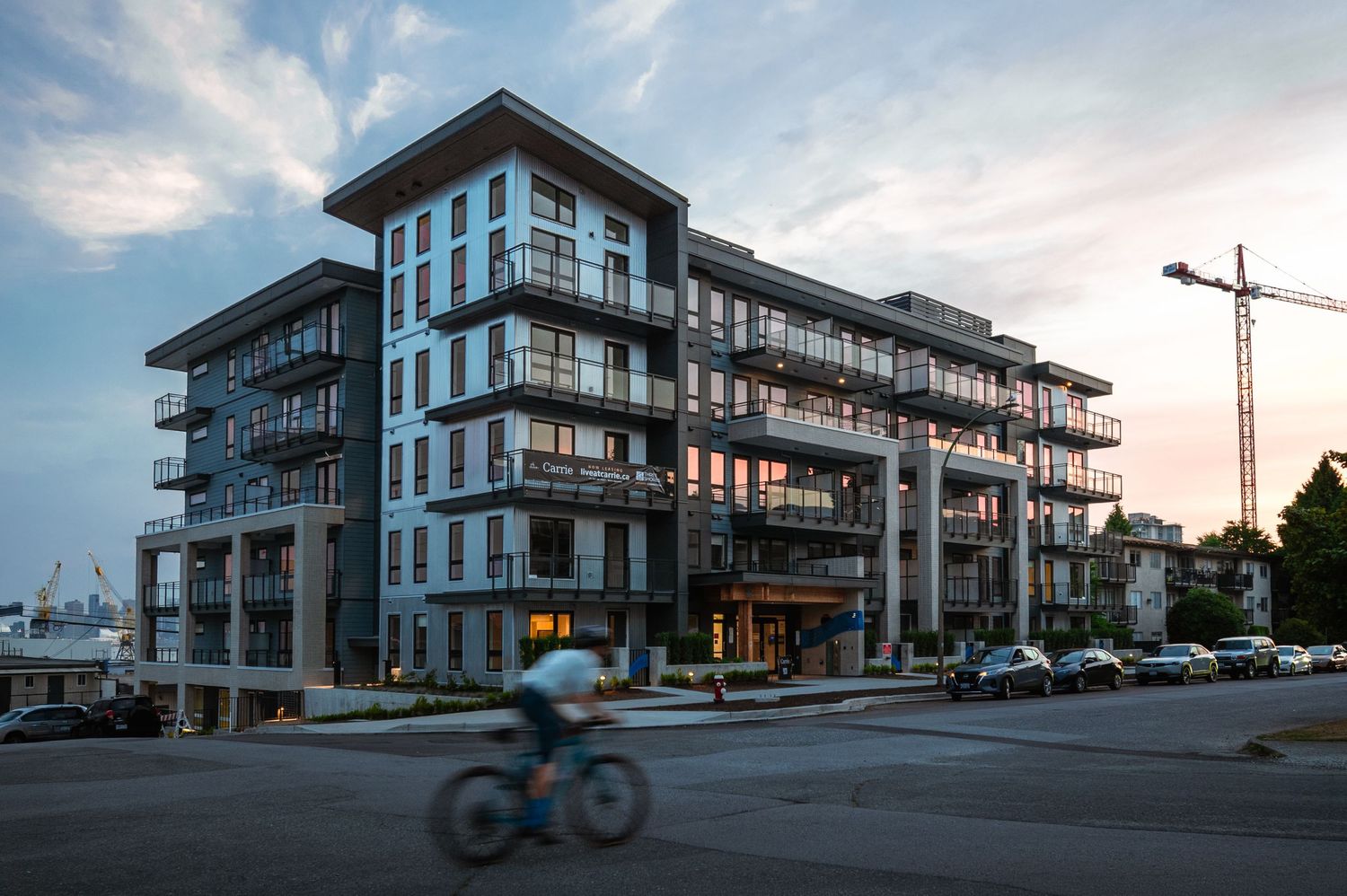 Modern multi-story residential building at dusk with a cyclist passing by and a construction crane in background.