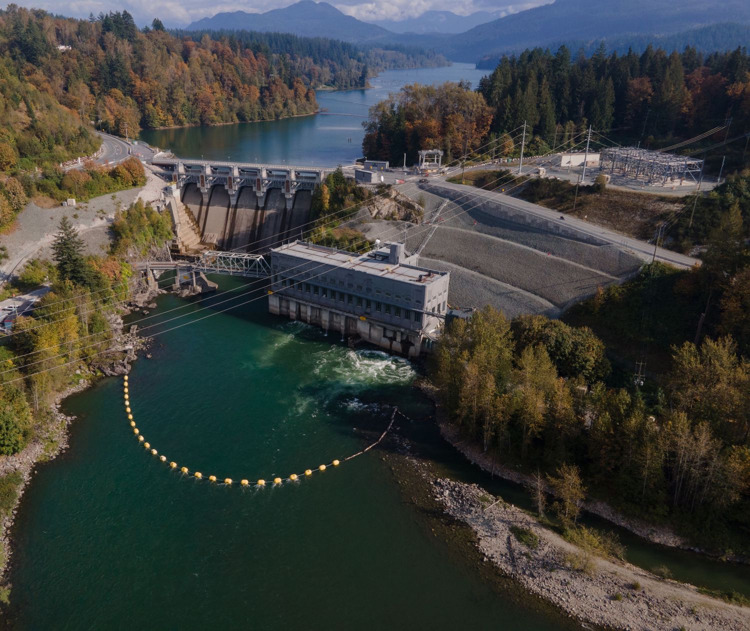 Aerial view of a hydroelectric dam and power station surrounded by forested mountains and a river.