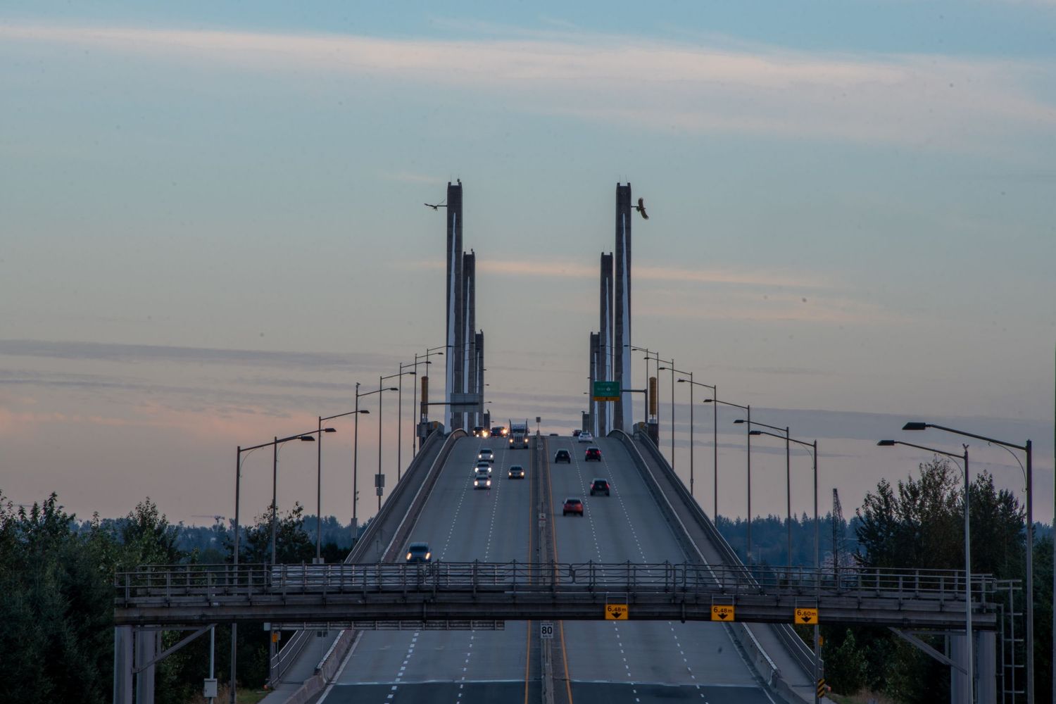 Cars drive over a cable-stayed bridge at dusk with towers rising against a cloudy sky.
