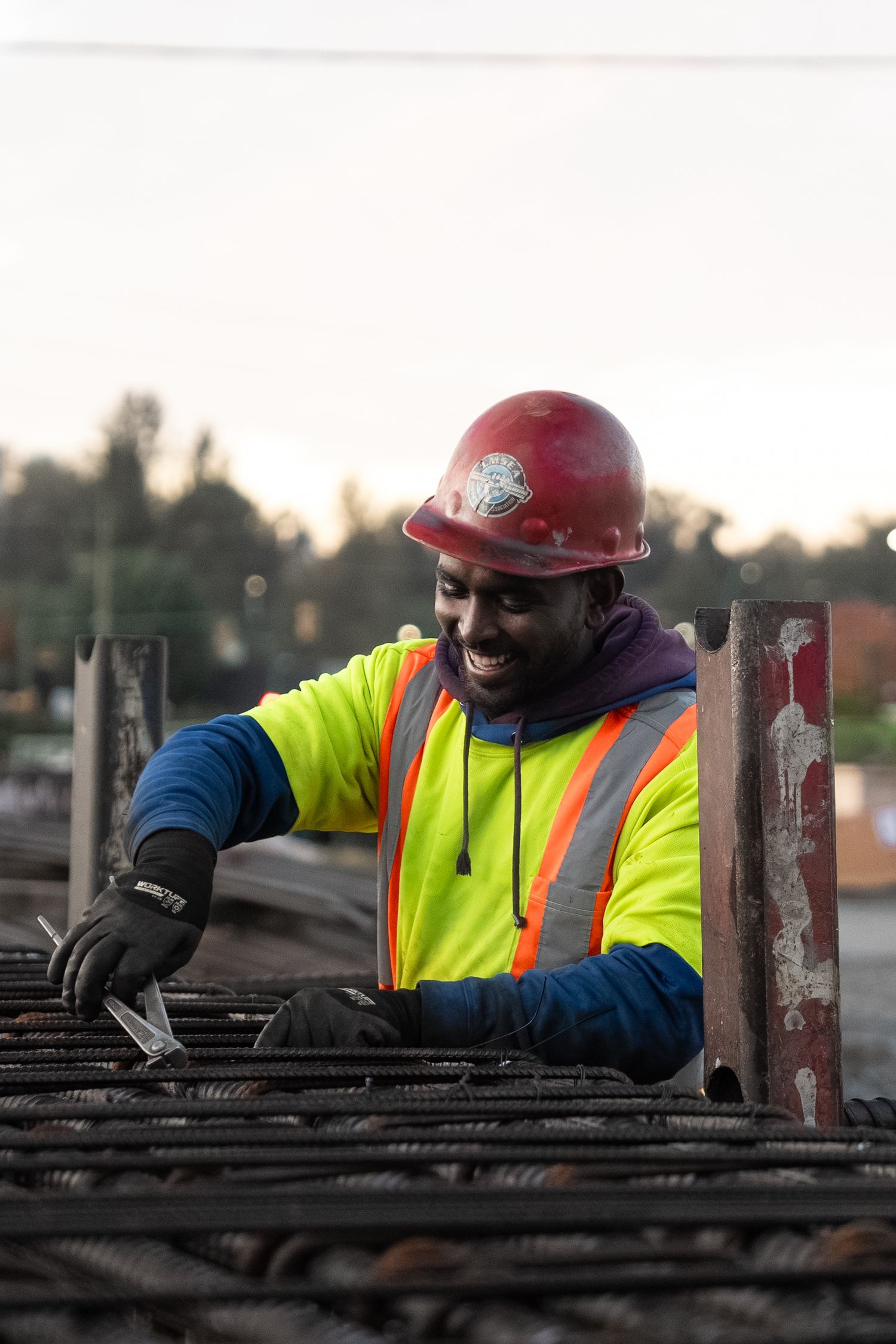 Smiling construction worker in yellow safety vest and red hard hat working with steel rebar at a job site.