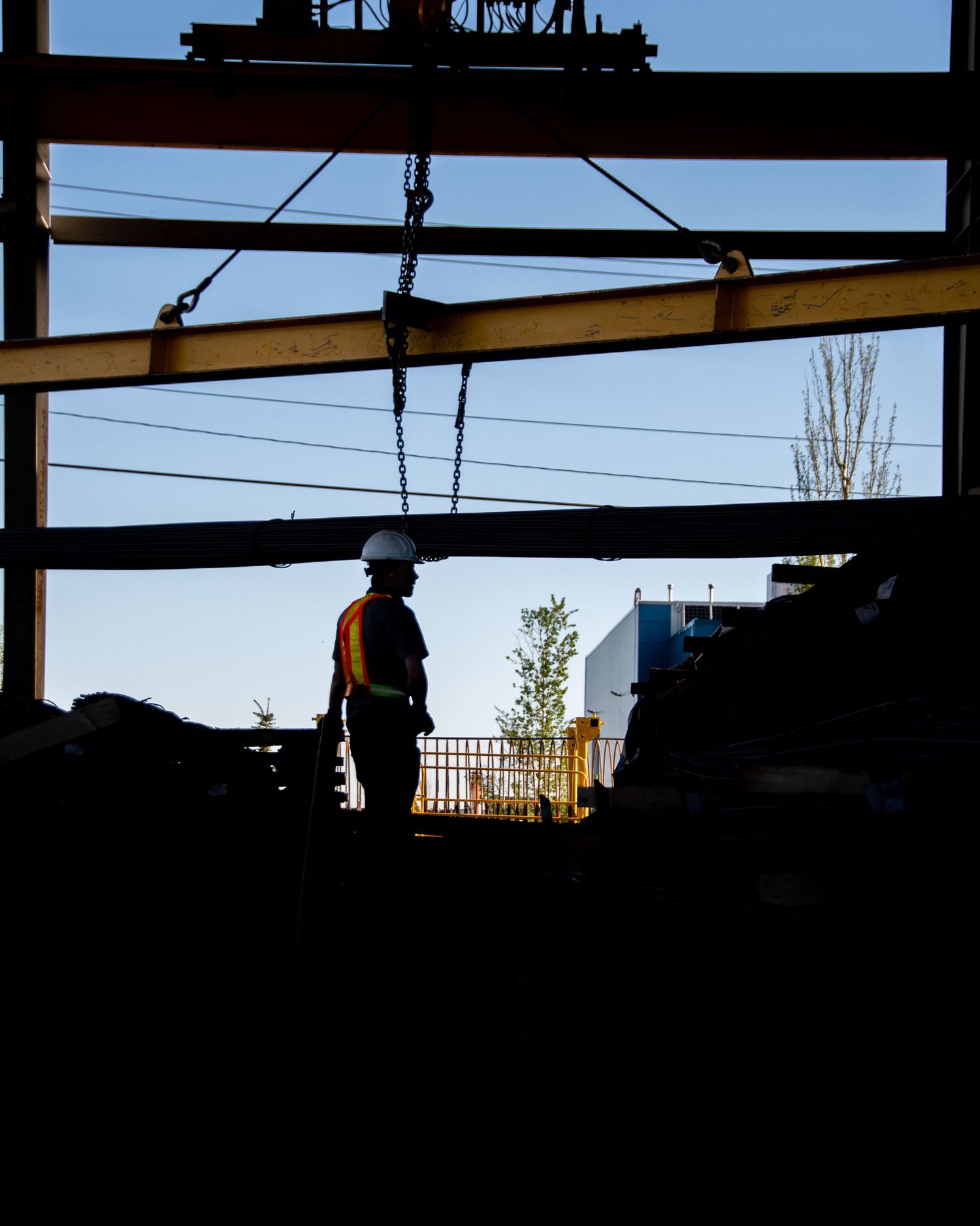 Silhouette of a construction worker in hard hat and safety vest standing beneath steel beams at a job site.