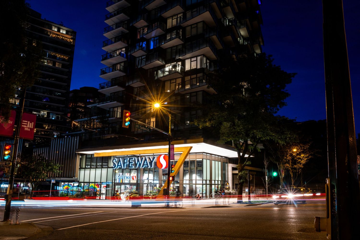 Safeway grocery store illuminated at night with light trails from passing cars at urban intersection.