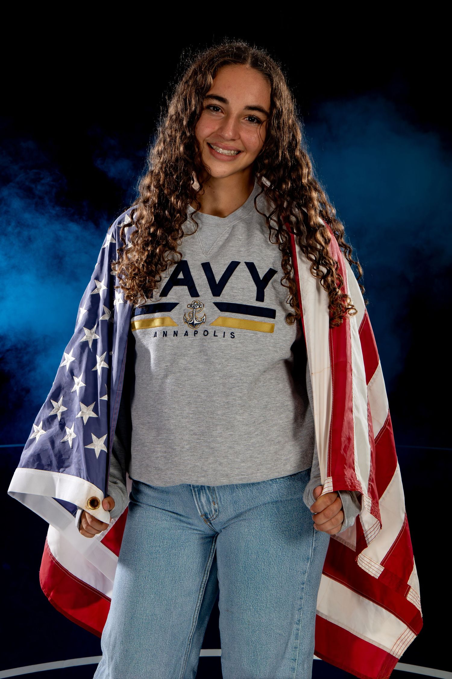 Smiling young woman wearing Navy Annapolis shirt draped with American flag in dramatic studio setting.