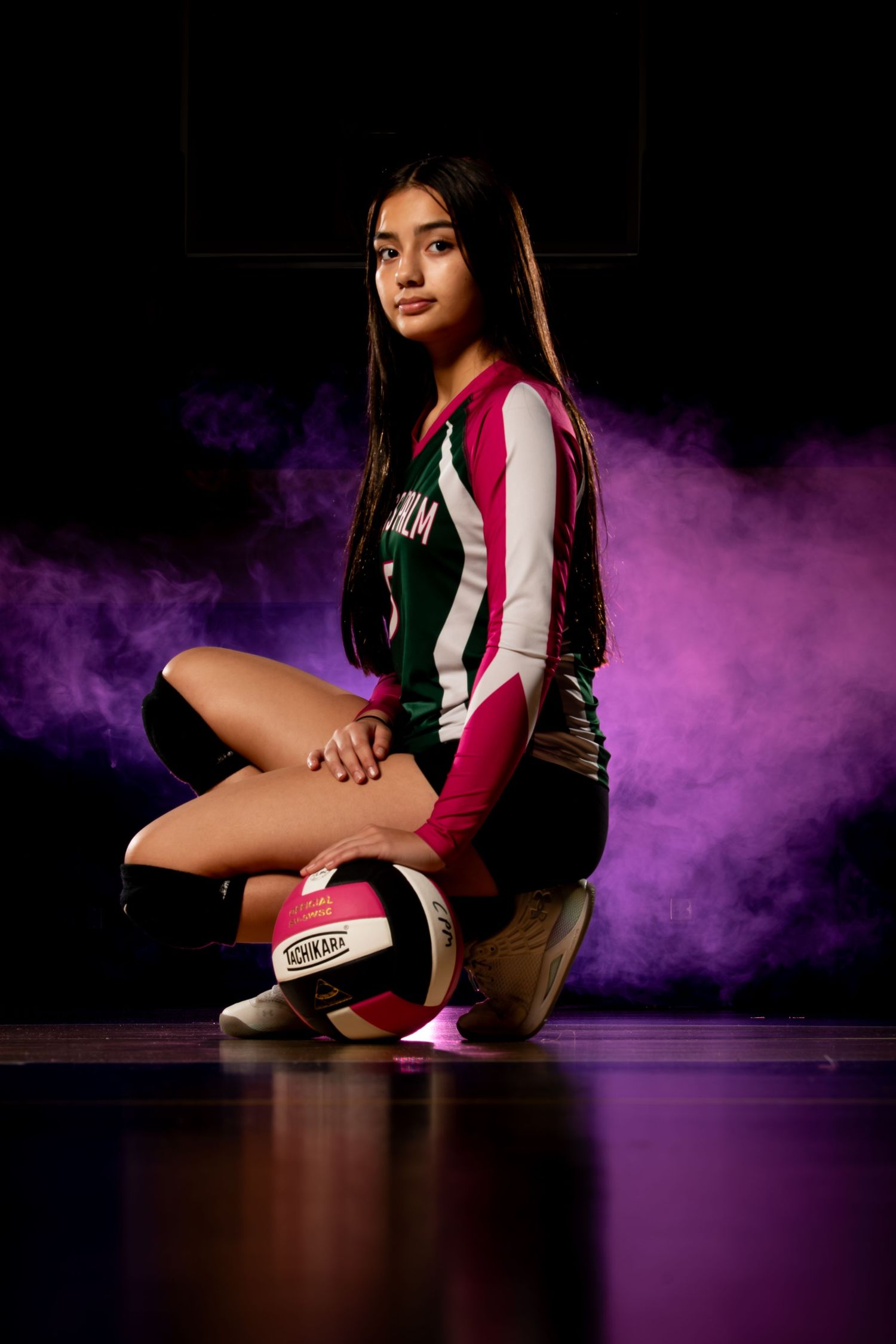 Female volleyball player in uniform poses with ball against dramatic purple smoke background in dark studio.