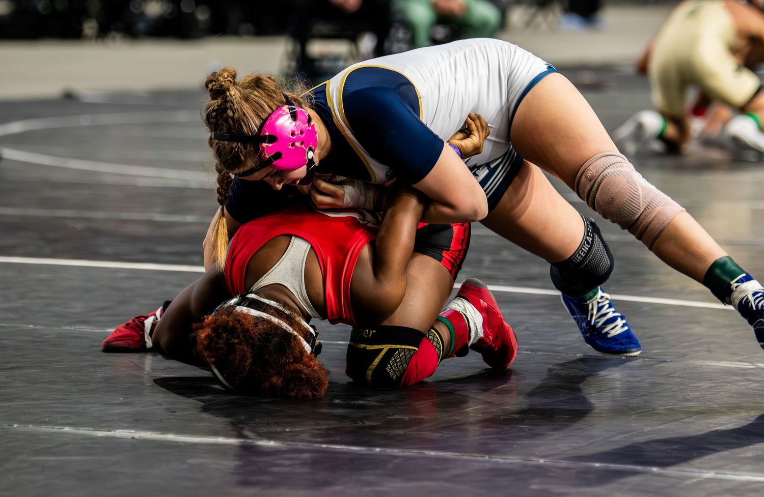 Two female wrestlers competing on mat, one in red pinned by opponent in blue uniform.