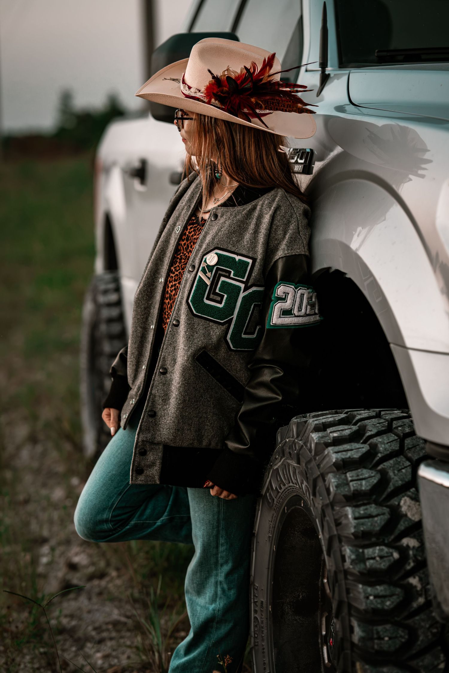 Young woman in varsity jacket, hat, and jeans leaning against white lifted truck outdoors.