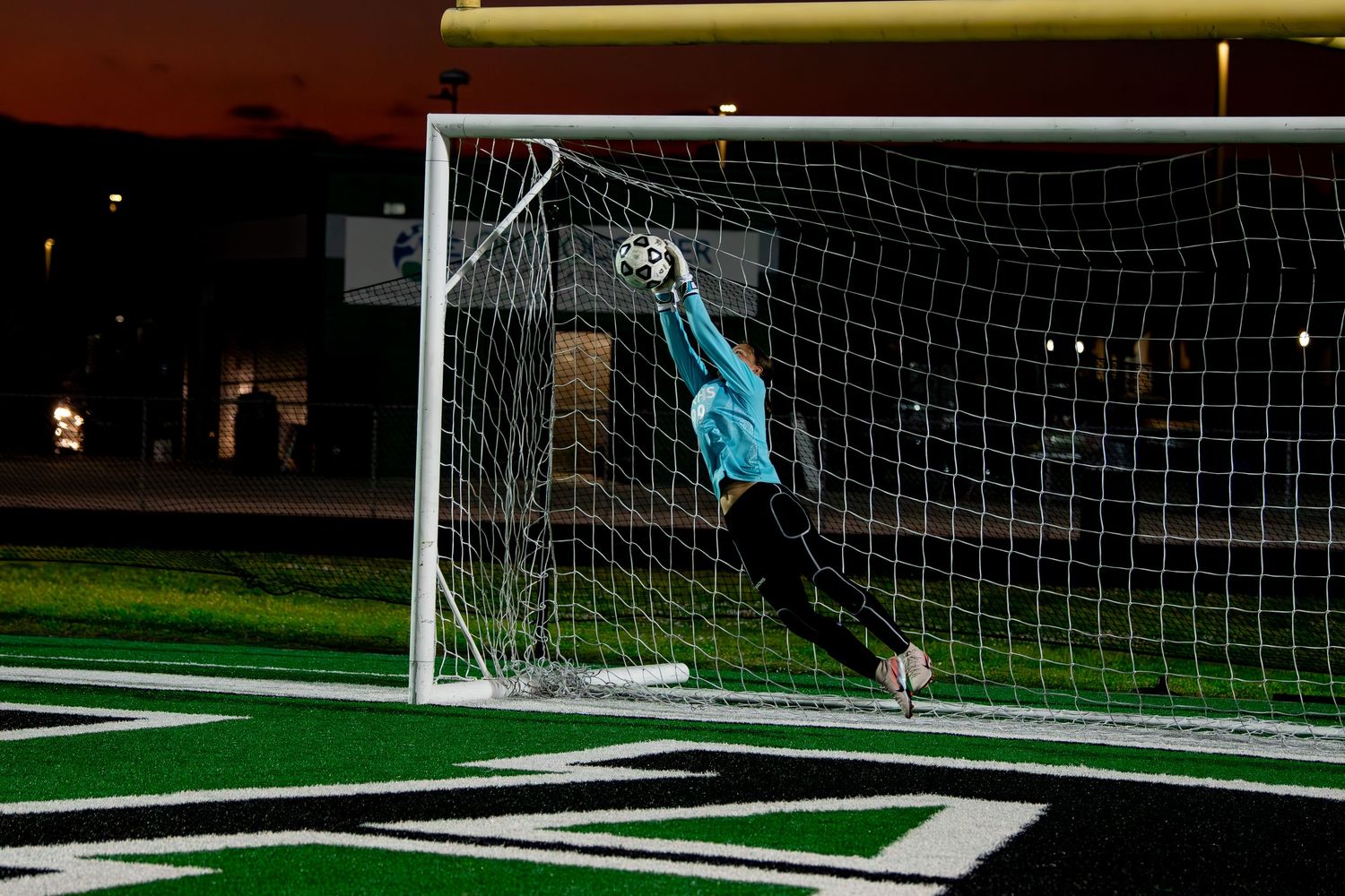 Soccer goalkeeper making a dramatic diving save at the goal during a night game on a lit turf field.