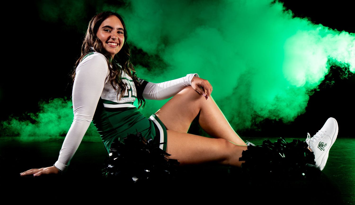 Smiling cheerleader in green uniform poses casually on floor surrounded by dramatic green smoke in athletic portrait.