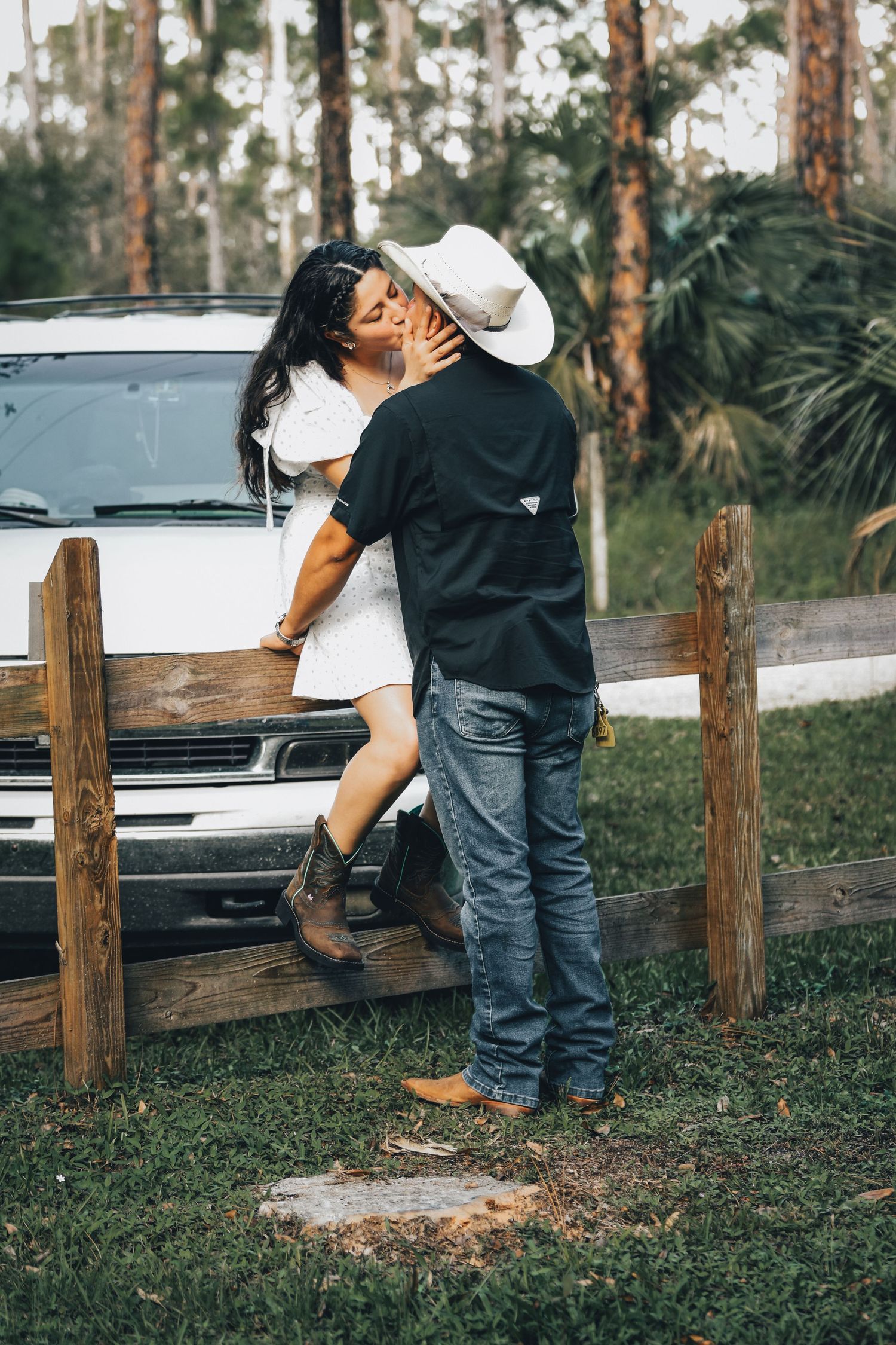 Couple sharing a romantic kiss by a wooden fence with a white truck and palm trees in background.