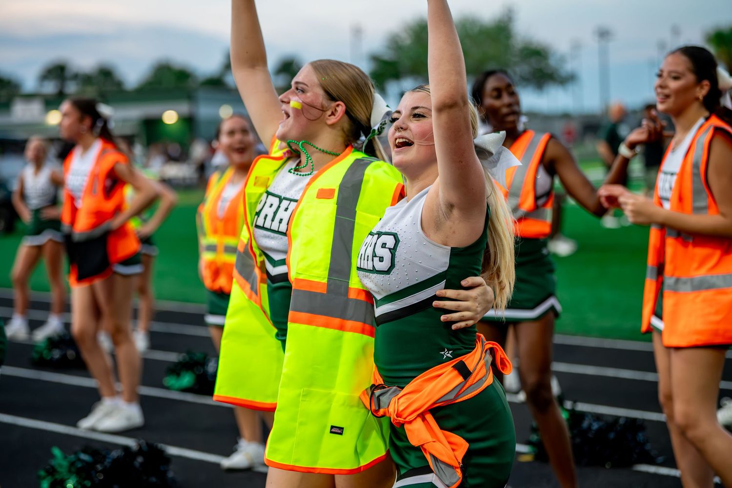 High school cheerleaders in green and white uniforms cheer enthusiastically at a football game.
