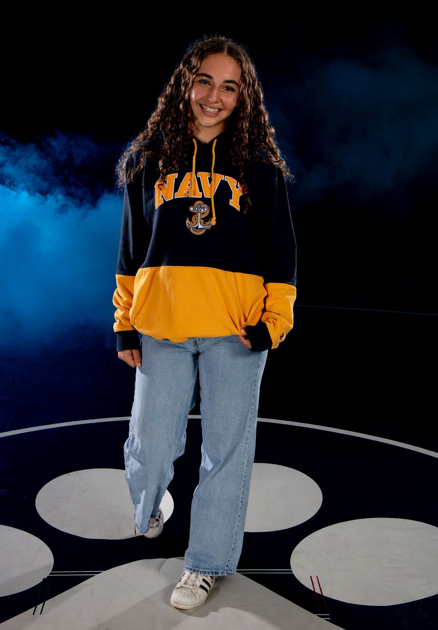 Young woman smiling in Navy hoodie and jeans posing on wrestling mat with dramatic blue smoke background.
