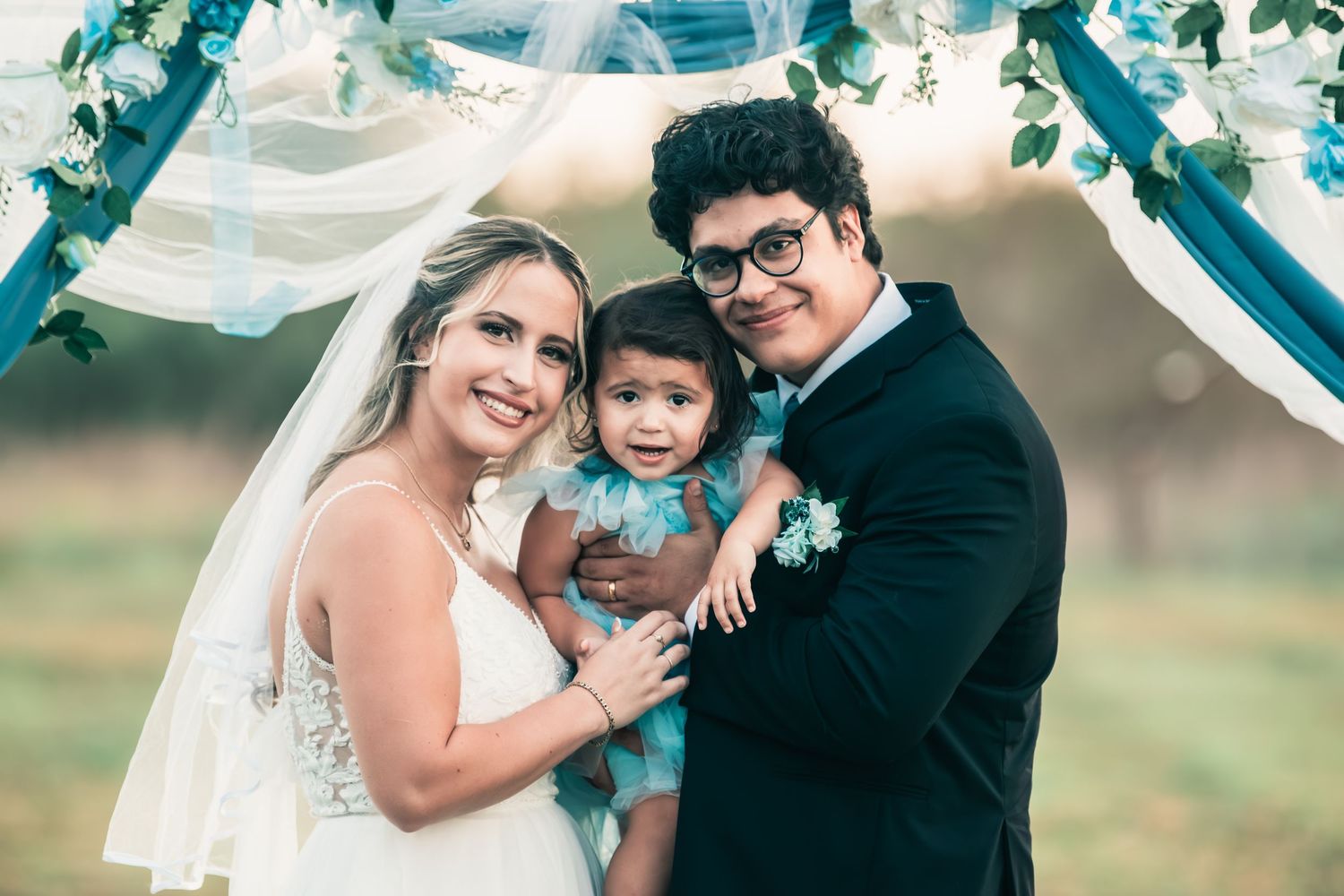 Couple in wedding attire holding a child beneath a floral arch.