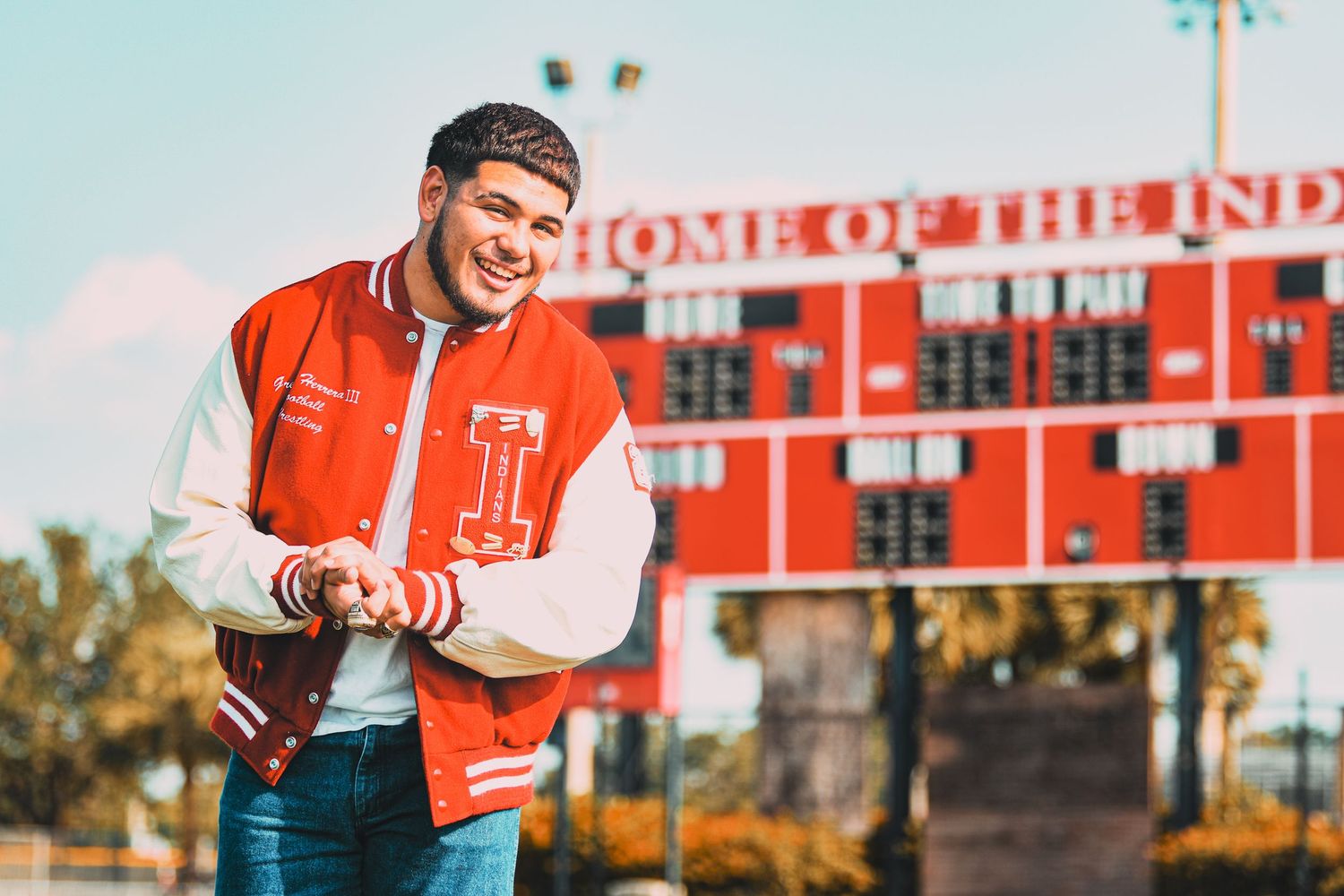 Smiling young man in red letterman jacket standing in front of high school scoreboard.