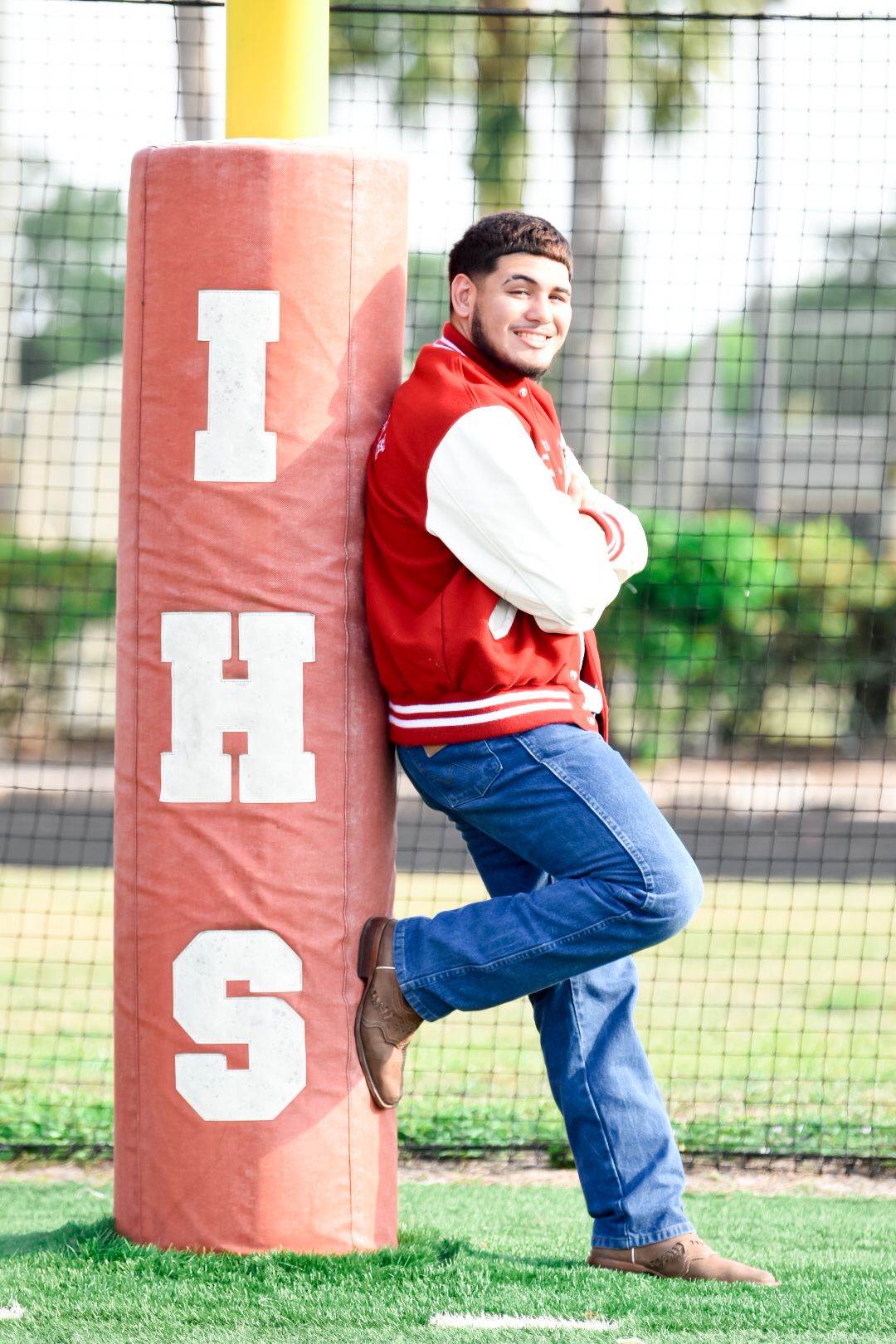 Young man in red letterman jacket leaning against IHS football field goal post, smiling with arms crossed.