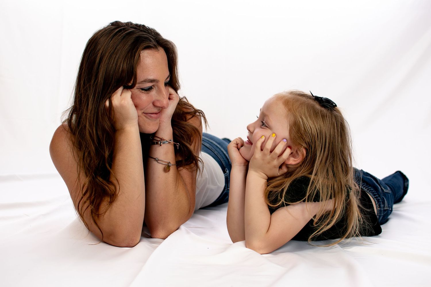 Smiling brunette woman and young blonde girl lying on white surface facing each other.
