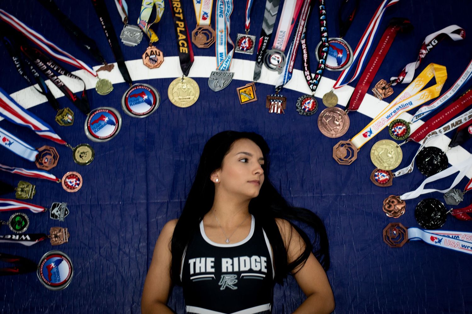 Young female athlete lying surrounded by circle of medals and trophies wearing The Ridge jersey.
