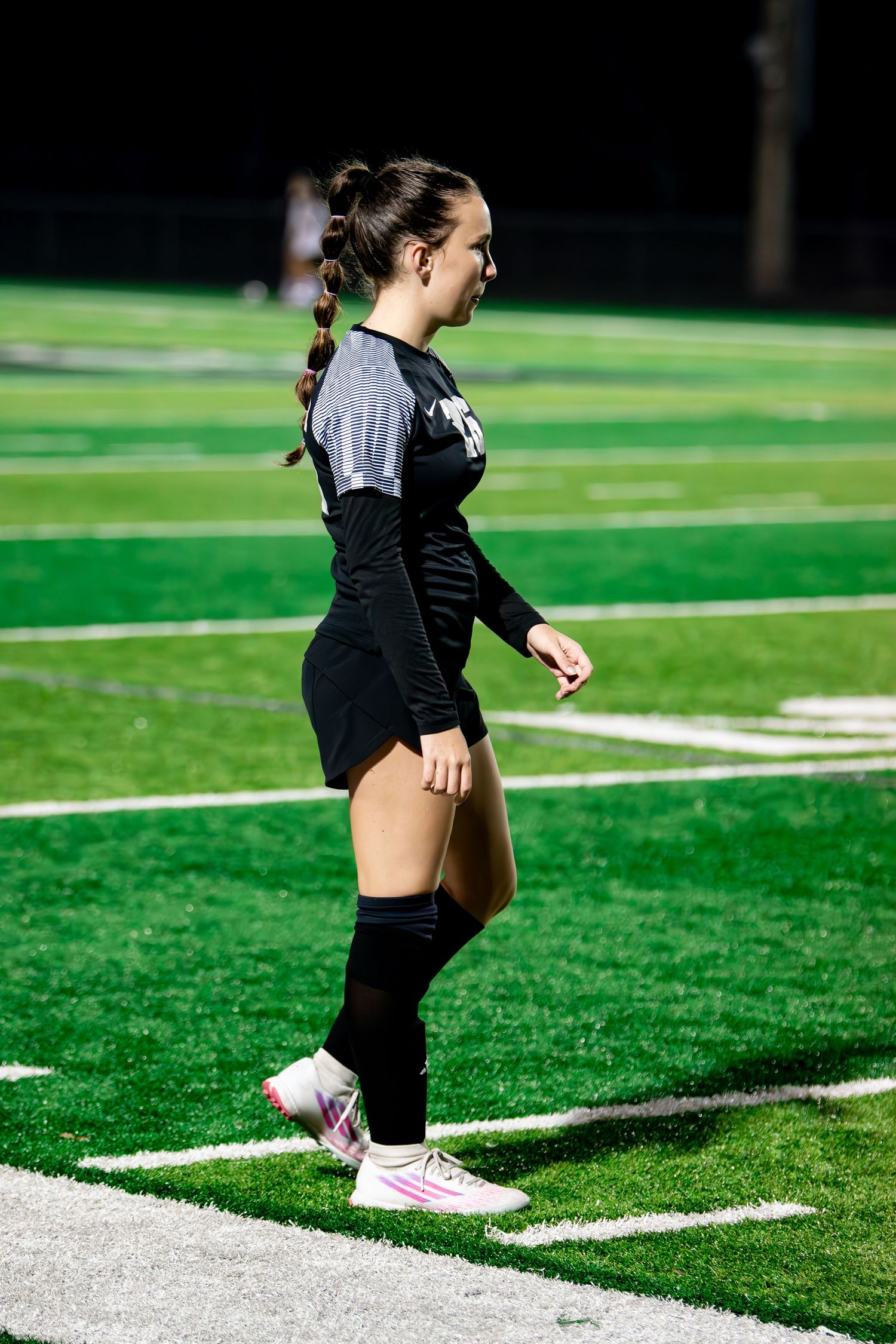 Female soccer player in black uniform walks along sideline during a night match.