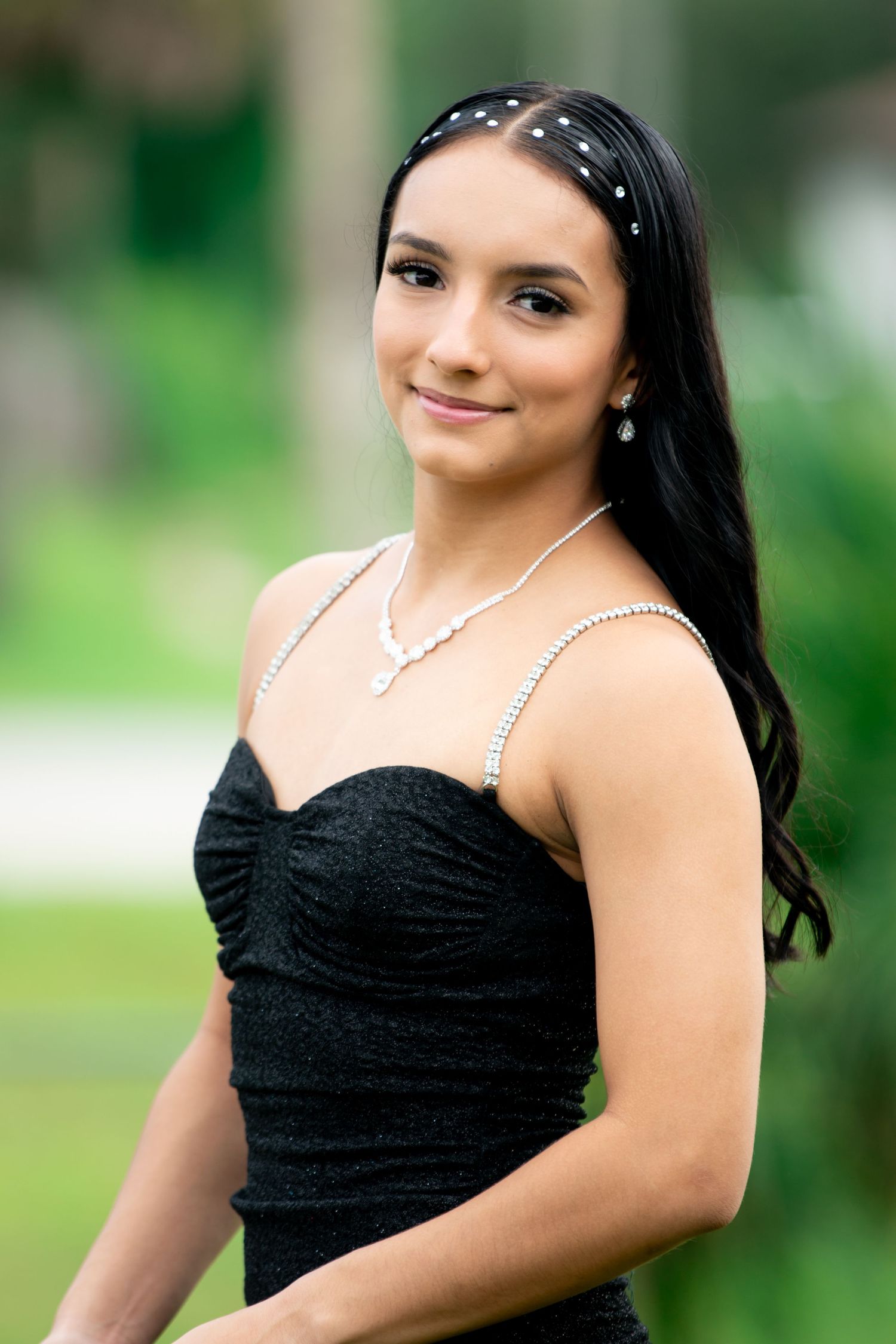 Young woman in black strapless dress with pearl necklace smiling outdoors.