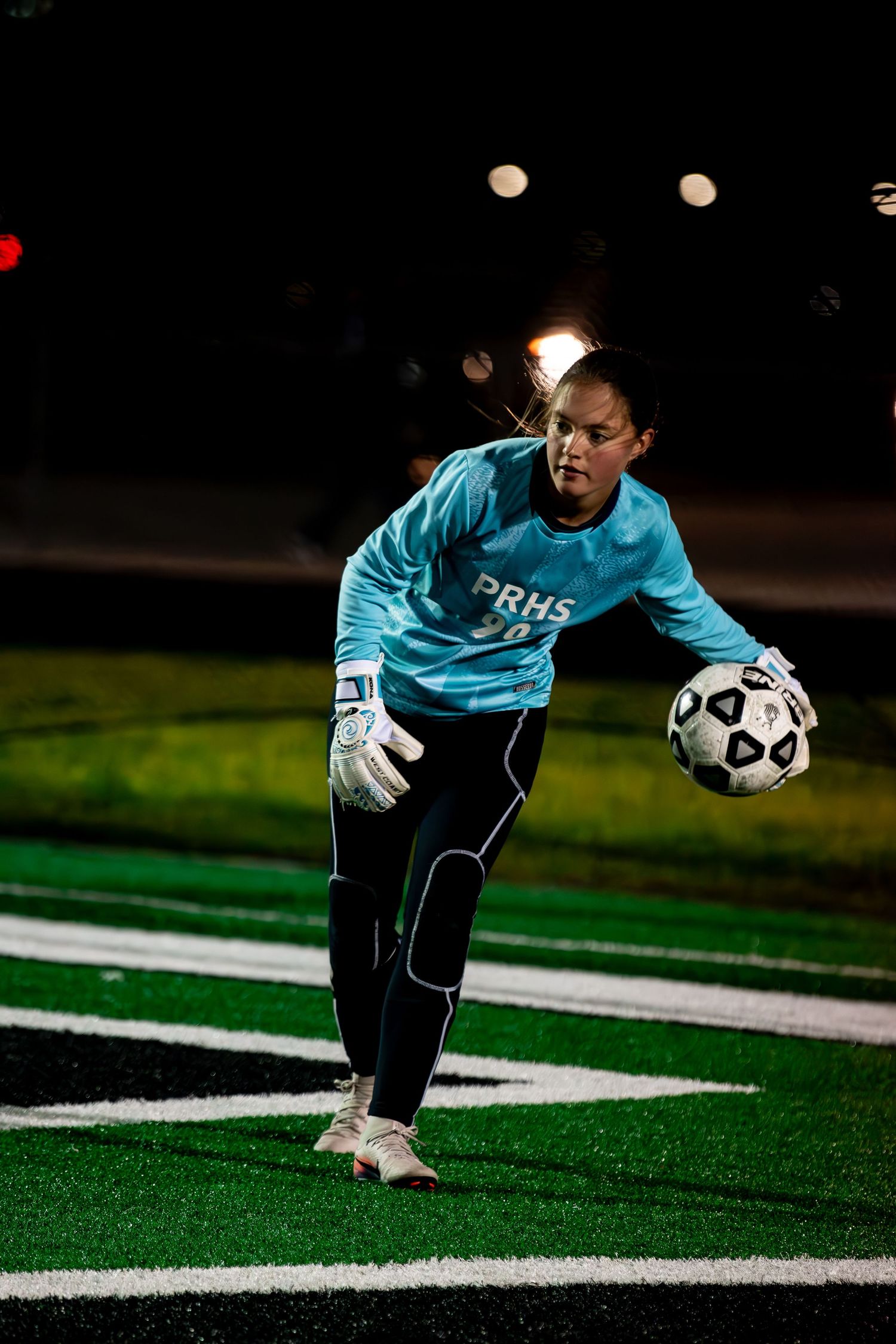 Female soccer goalkeeper in teal PRHS jersey prepares to throw ball during night game.