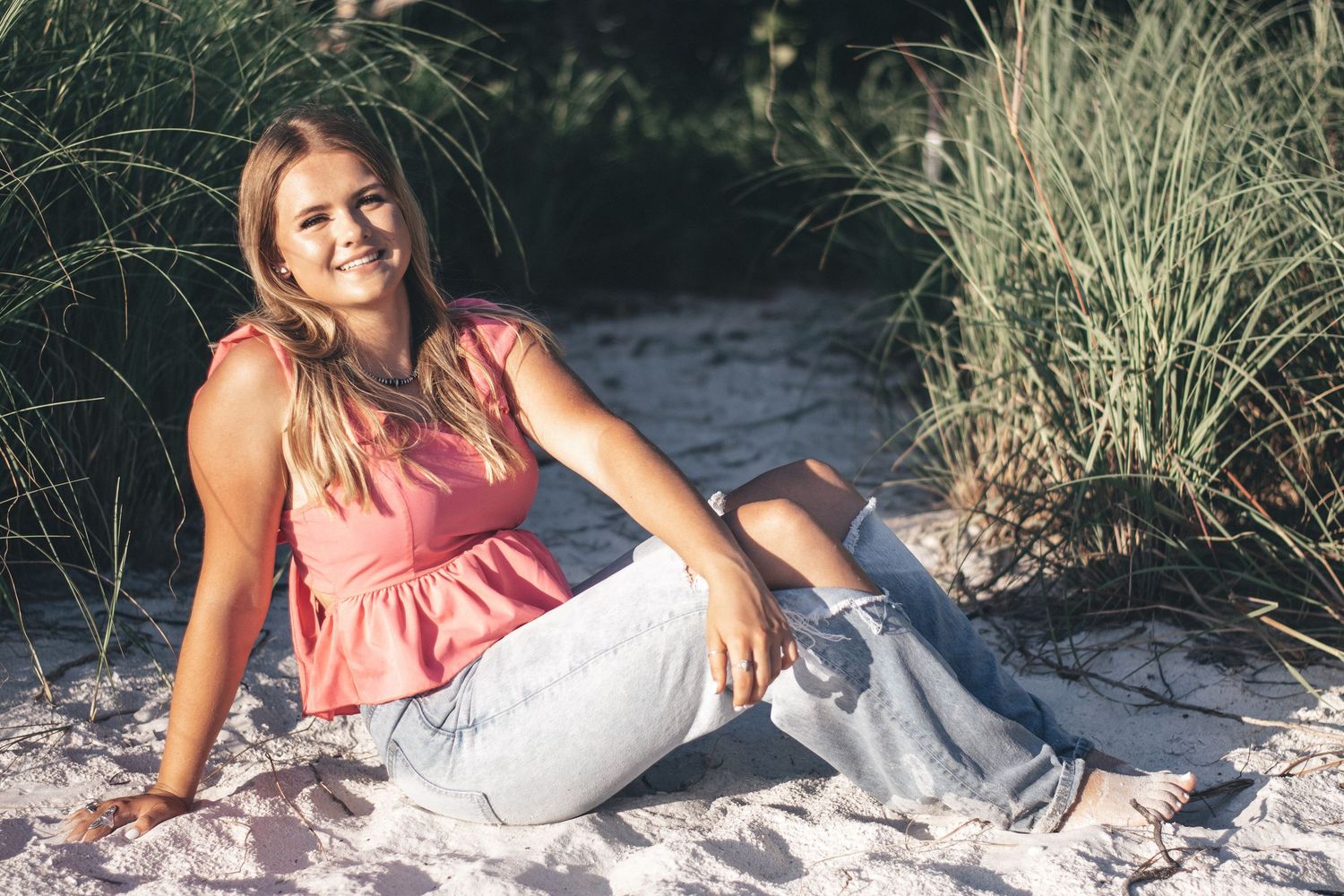 Smiling young woman in pink top and ripped jeans sitting on sandy beach dunes.