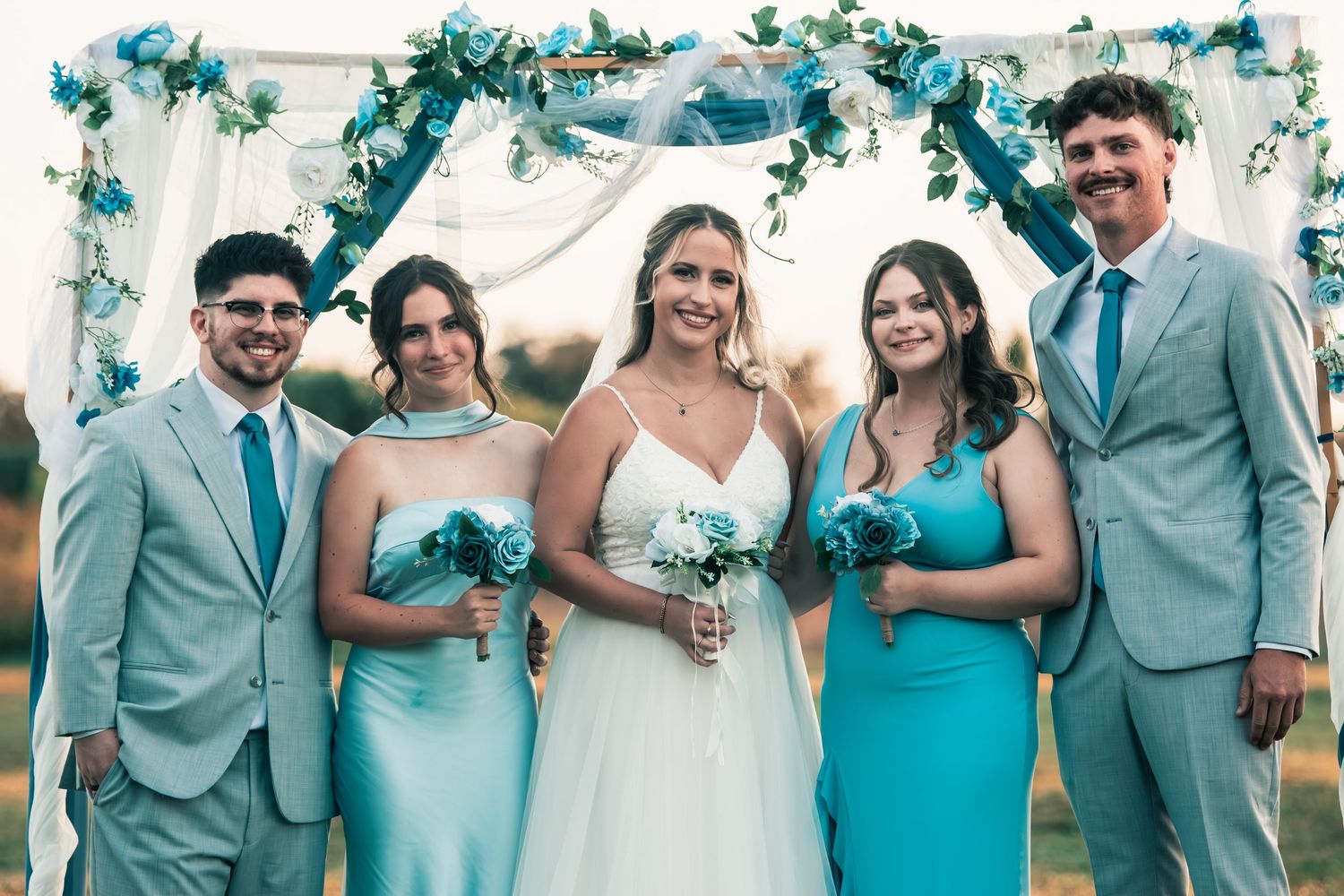 Bride in white gown posing with bridesmaids in teal dresses and groomsmen under blue floral arch.