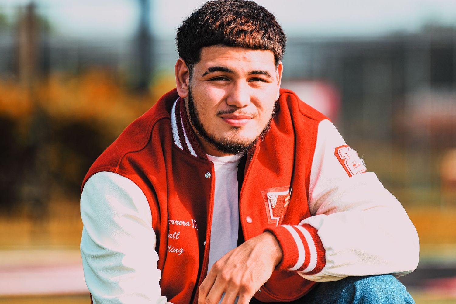 Young man in red and white letterman jacket smiling during outdoor senior portrait session.