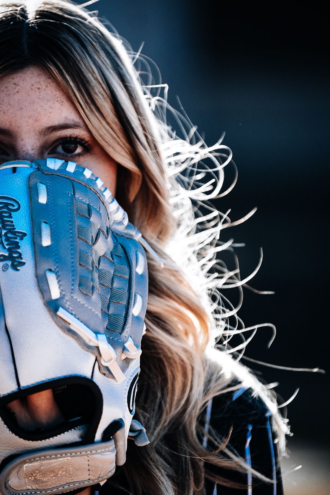 Close-up of blonde softball player holding blue Rawlings glove up to her face on dark background.