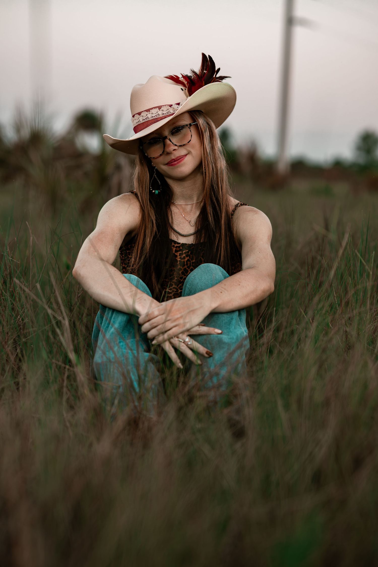Young woman in feathered cowboy hat and glasses sitting in tall grass field wearing teal pants.