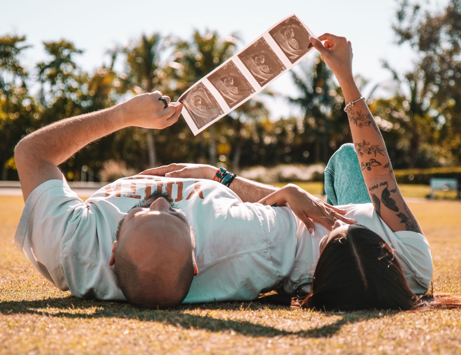 Couple lying on grass holding up ultrasound photos to announce their pregnancy outdoors.