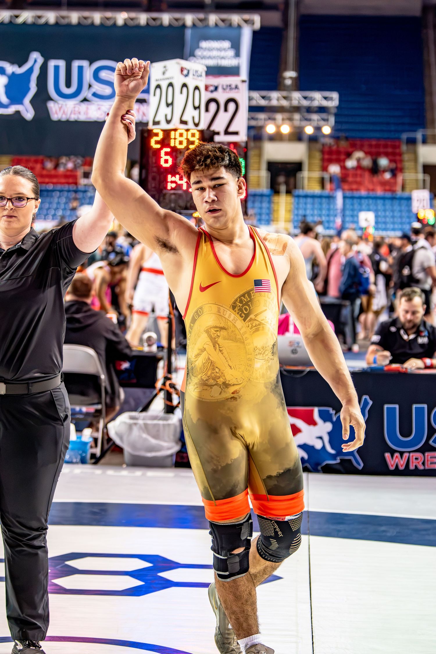 Young male wrestler in gold singlet has his arm raised in victory at a USA Wrestling championship event.