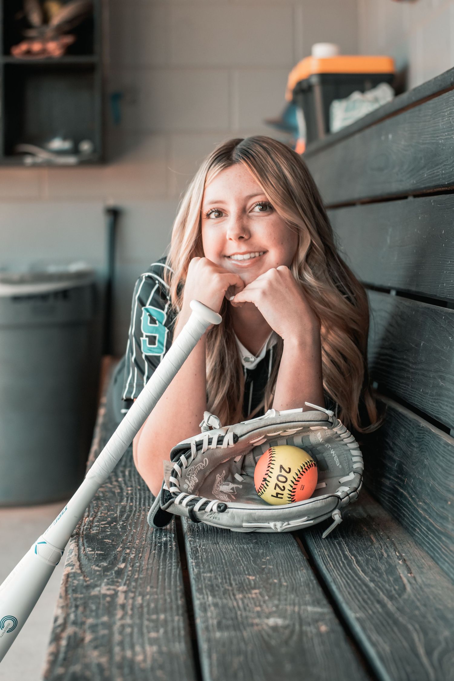 Smiling blonde softball player rests on dugout bench with glove, ball, and bat in uniform.