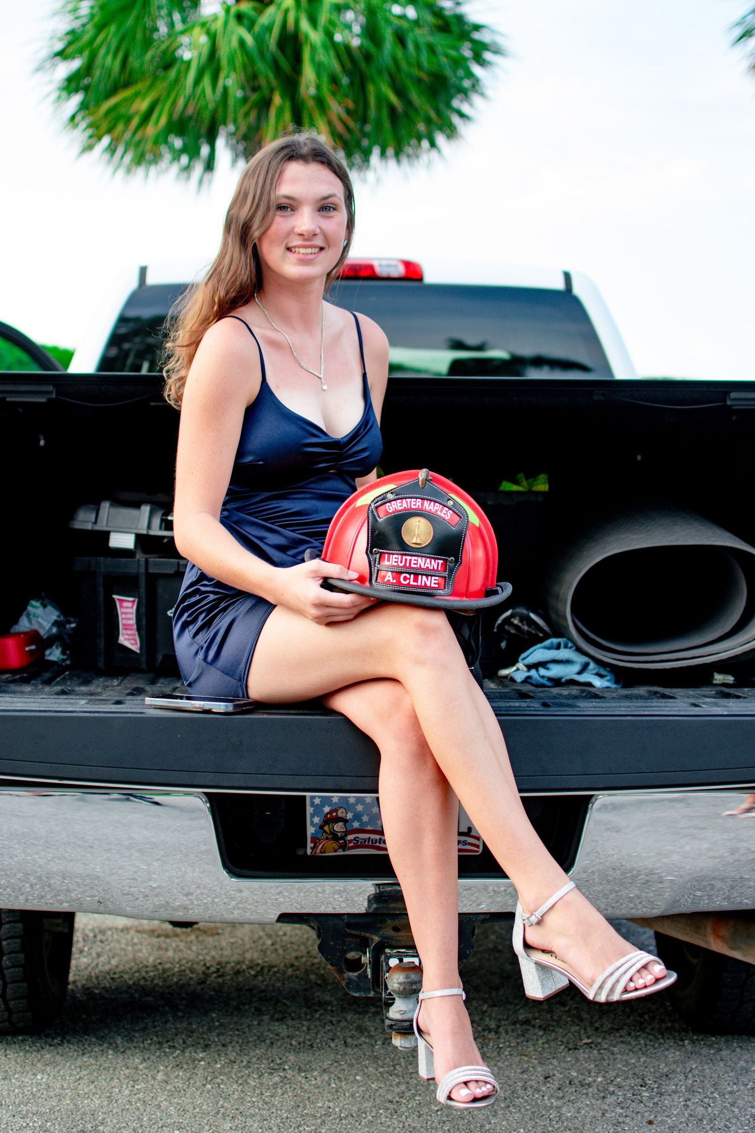 Young woman in blue dress holding firefighter helmet sitting on truck tailgate.
