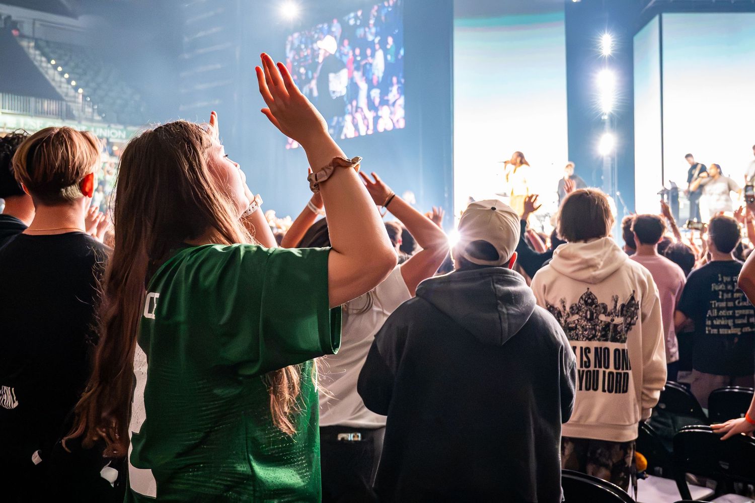Young woman in green shirt raises hands in worship at a large Christian concert event.