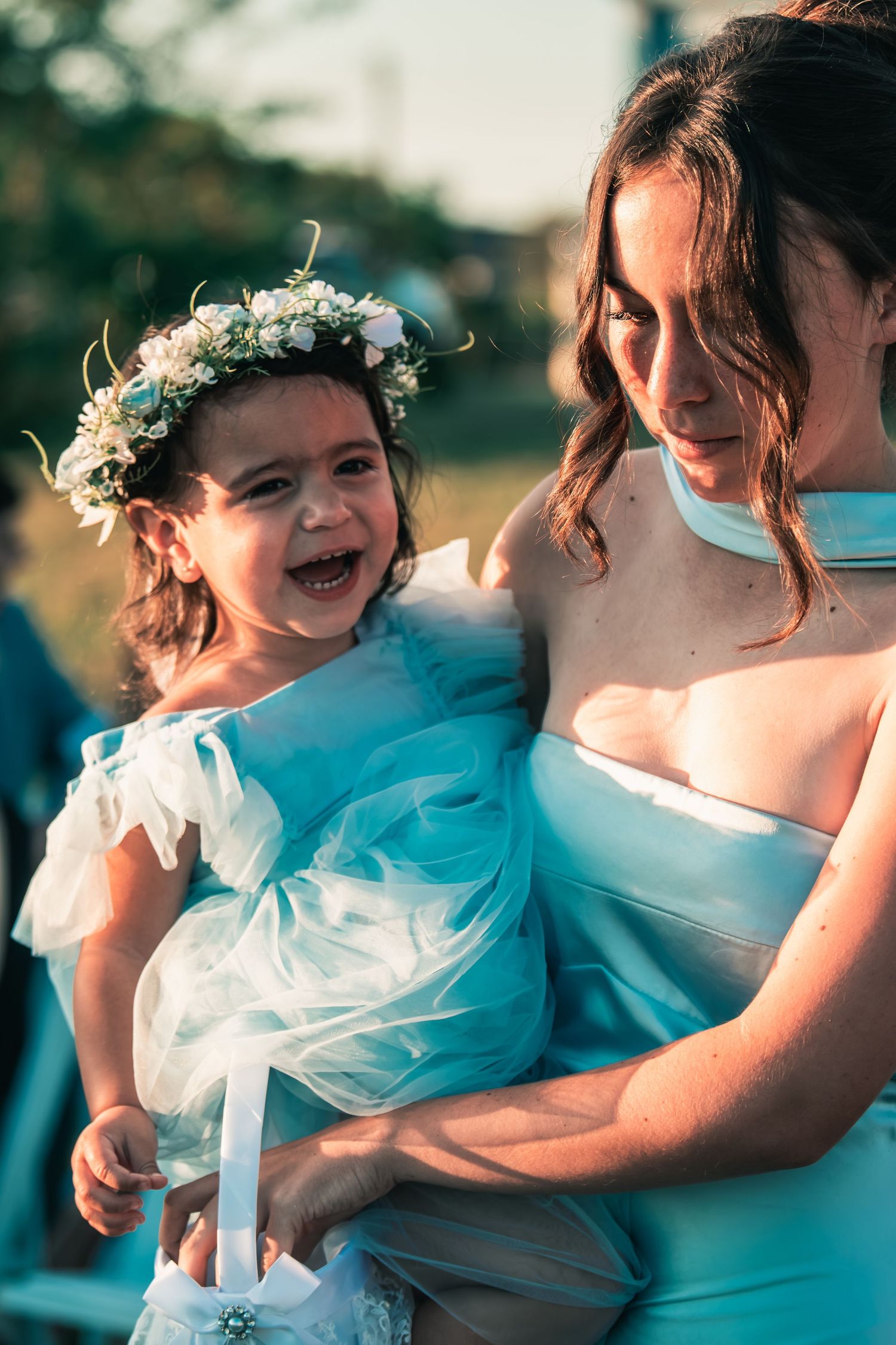 Bridesmaid in light blue dress holding laughing toddler flower girl wearing white floral crown.