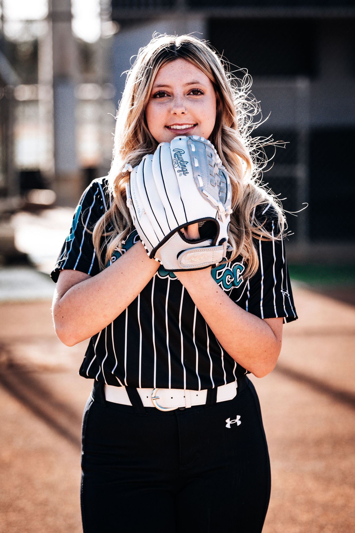 Blonde female softball player in pinstripe jersey holds white glove up to her face outdoors.