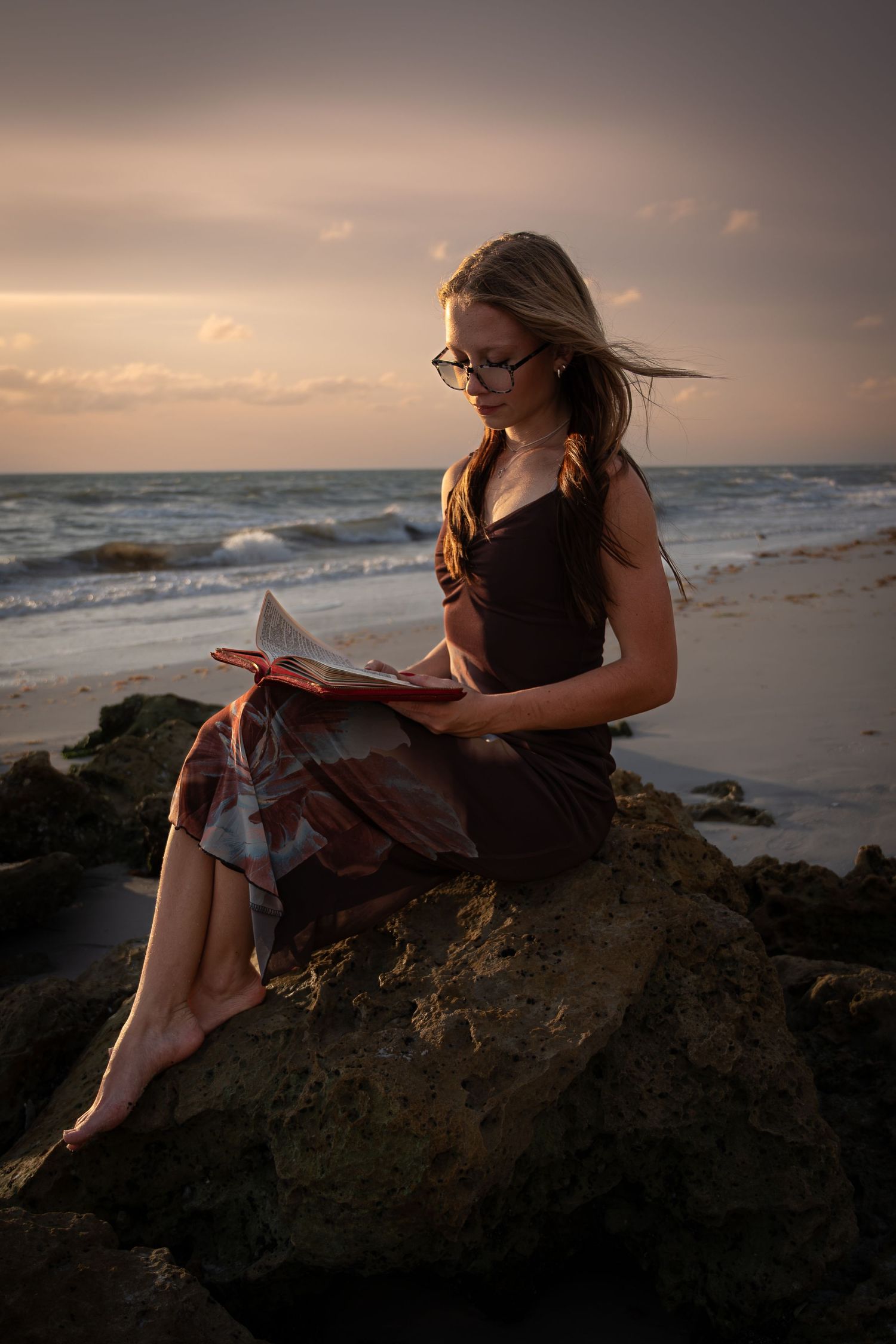 Young woman with glasses reading a book while sitting on rocks at a scenic beach during golden hour sunset.